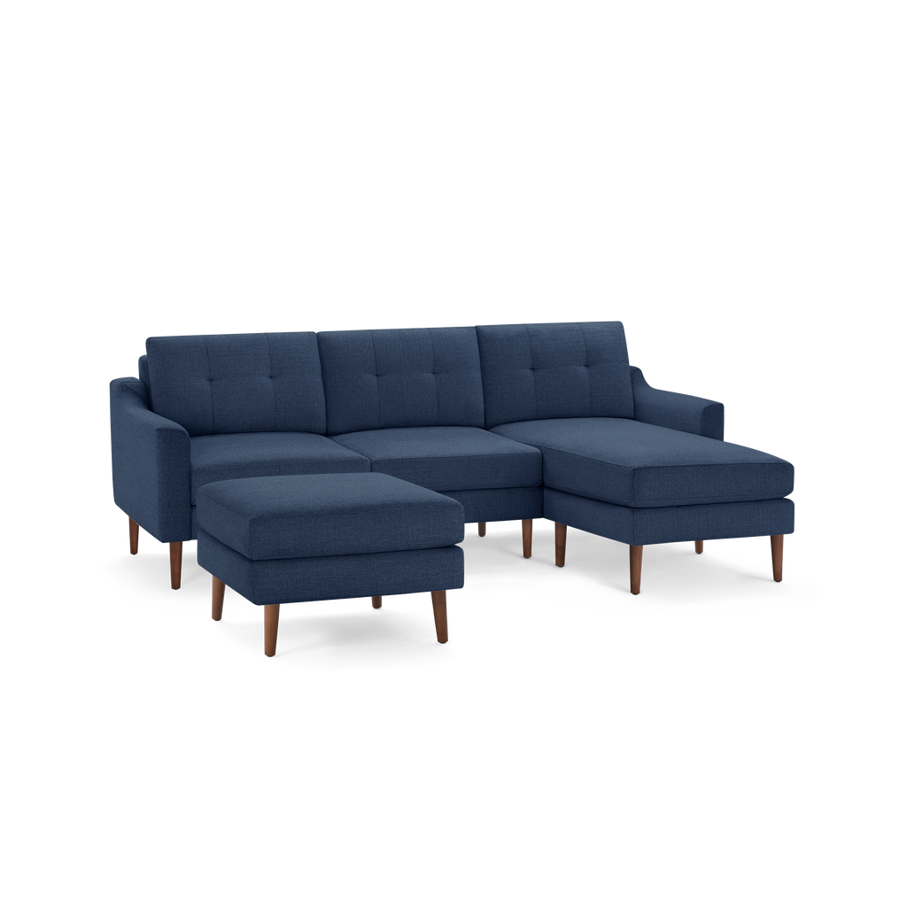 Modern navy blue three-seater sectional sofa with tufted cushions, wooden legs, and a matching ottoman on a transparent background.