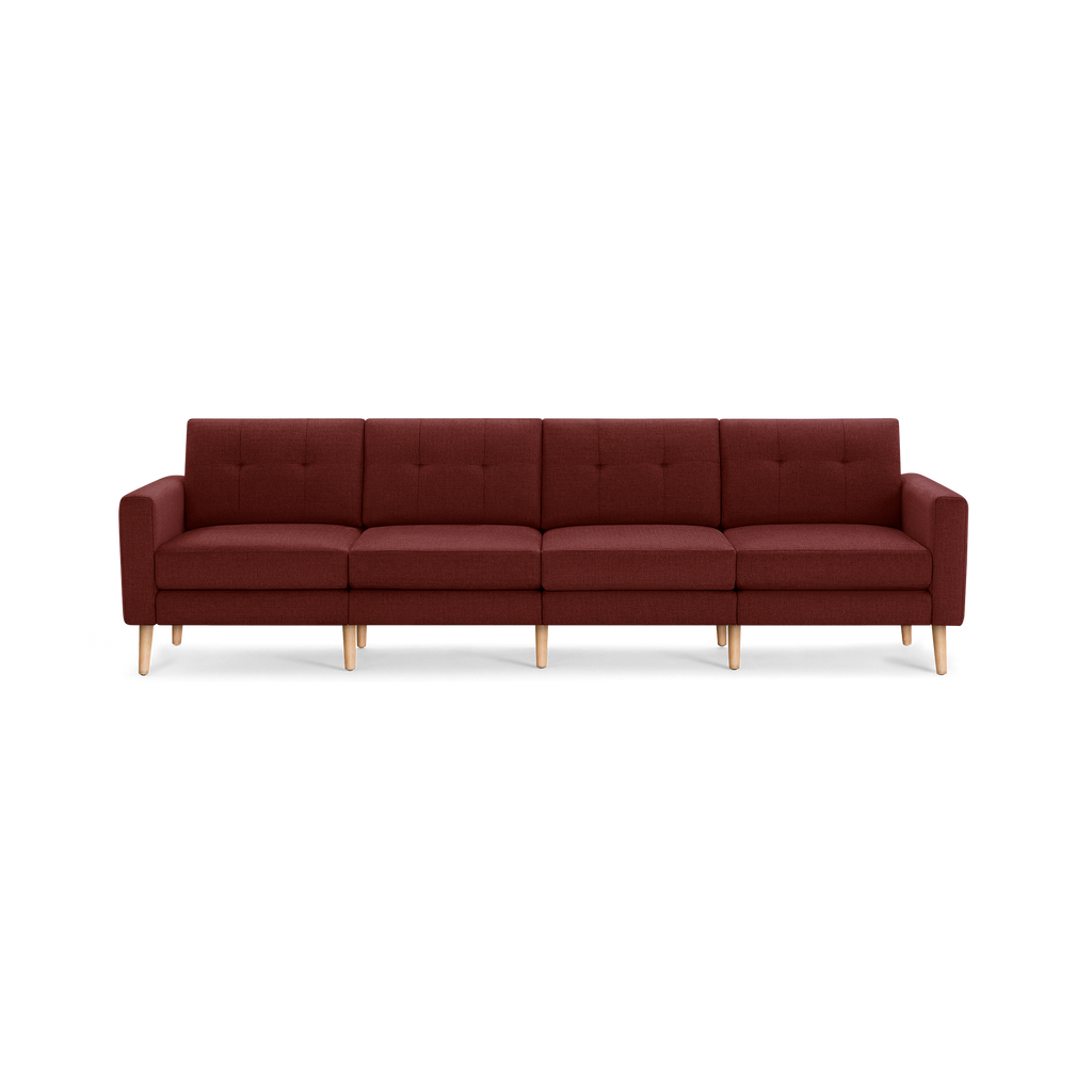 Modern long four-seater sofa with deep red fabric upholstery, tufted backrest cushions, and light wooden legs on a black background.