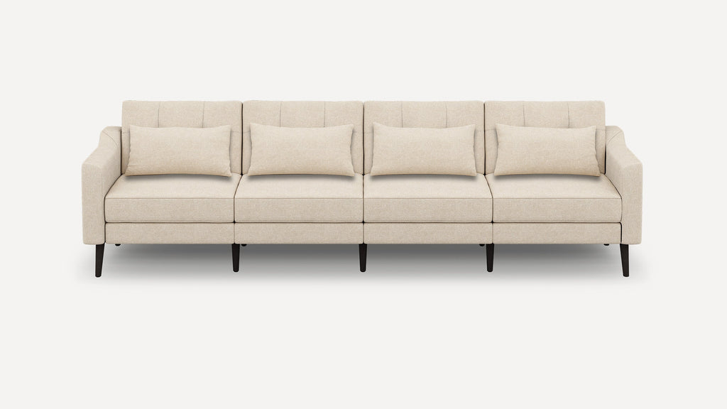 Beige modern four-seater sofa with clean lines, tufted back cushions, matching lumbar pillows, and dark wooden legs against white background.