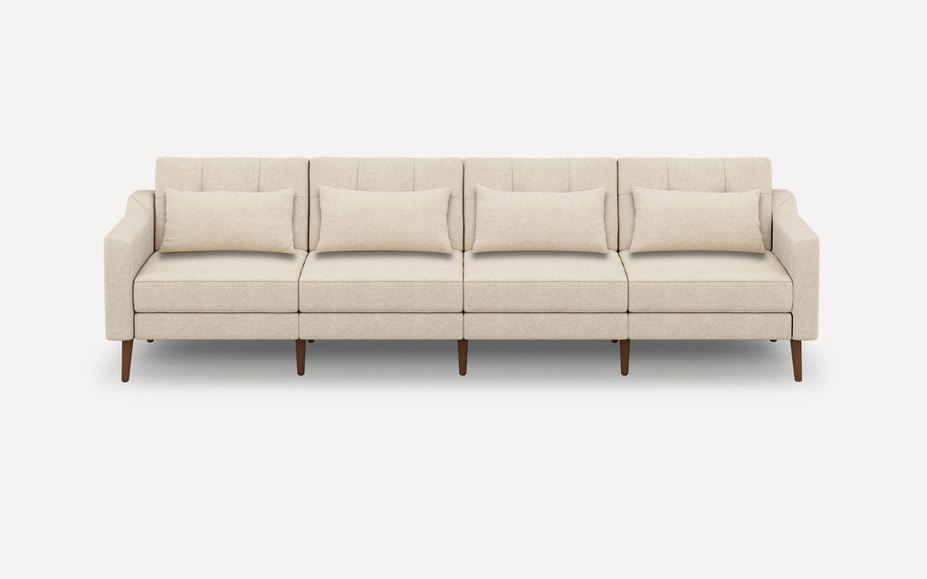 Modern four-seater beige fabric sofa with slim wooden legs, tufted back cushions, and matching rectangular throw pillows.