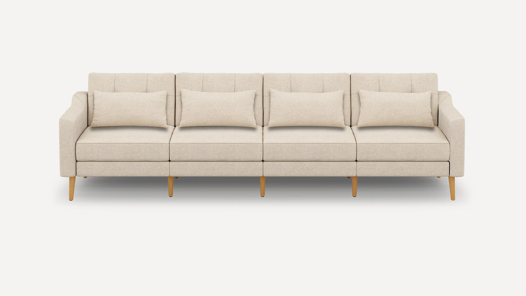 Four-seater beige fabric sofa with wooden legs, straight armrests, tufted back cushions, and four matching lumbar pillows.