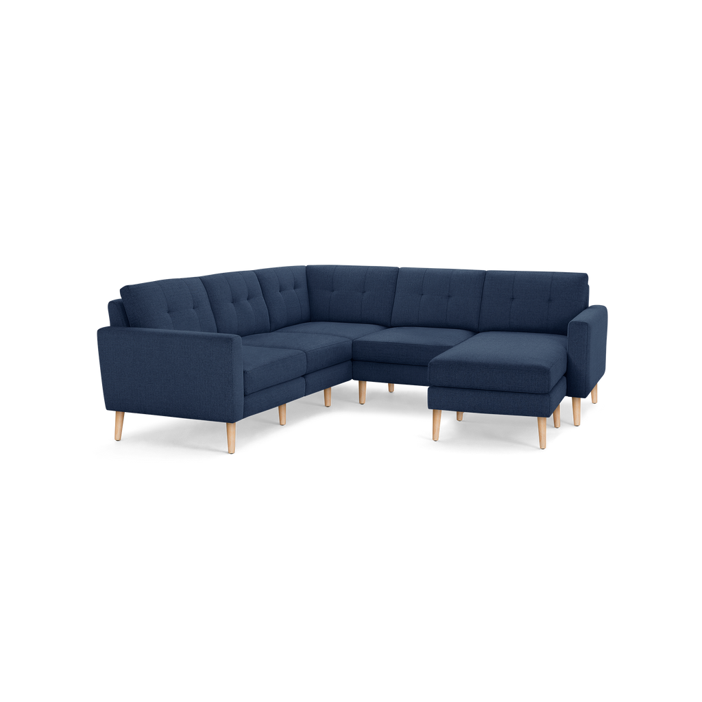 Modern L-shaped sectional sofa with dark blue fabric upholstery and light wooden legs, featuring tufted back cushions and a chaise lounge on the right side.