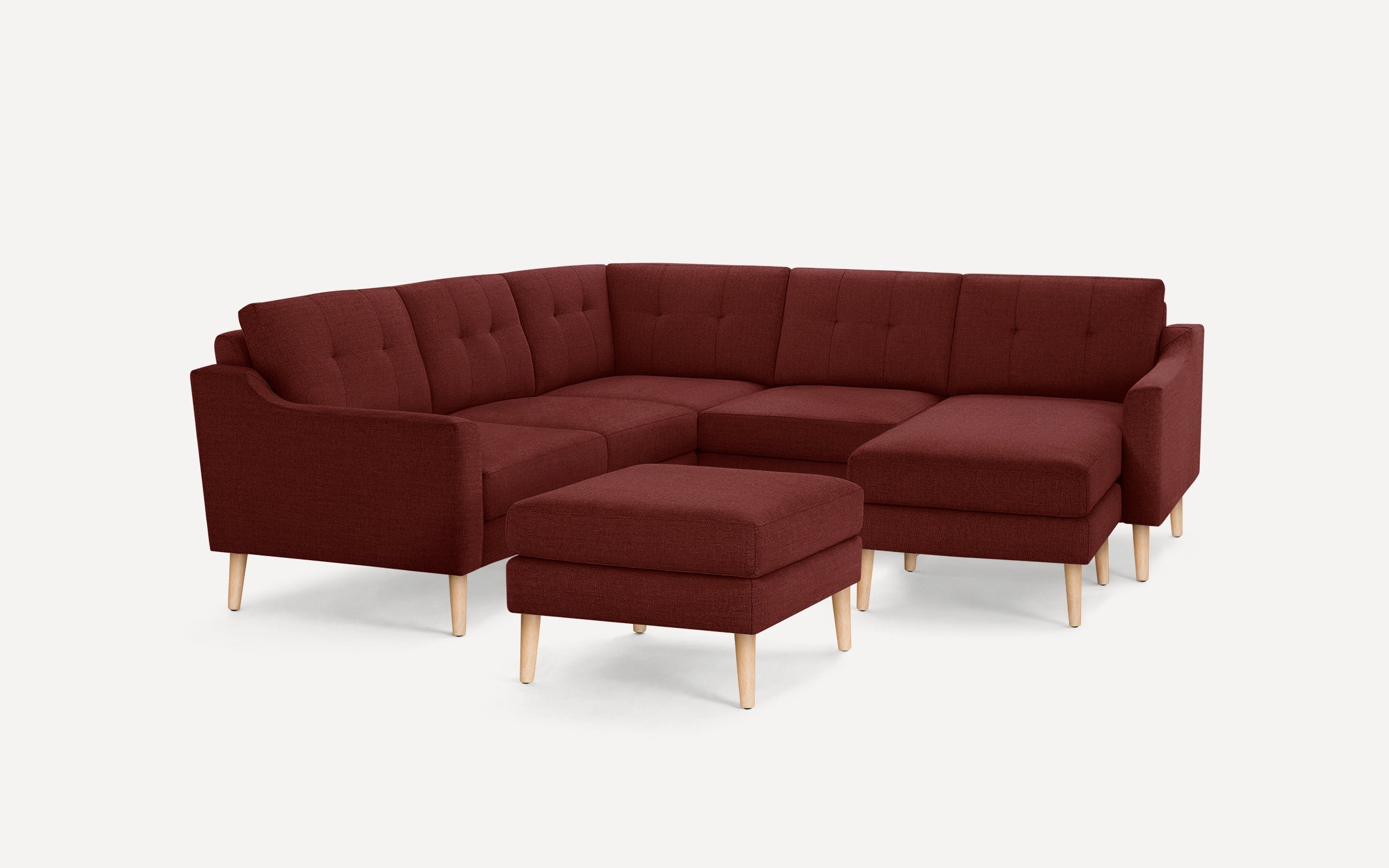 Modern L-shaped sectional sofa in deep burgundy fabric with tufted back cushions and light wooden legs, including a matching ottoman.
