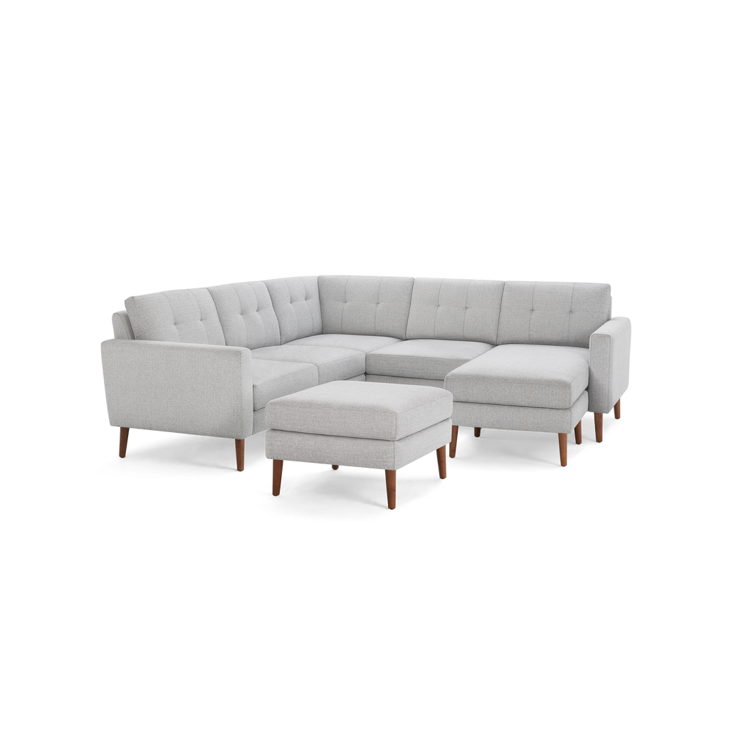 Light gray L-shaped sectional sofa with tufted back cushions, wooden legs, and matching ottoman on a black background.