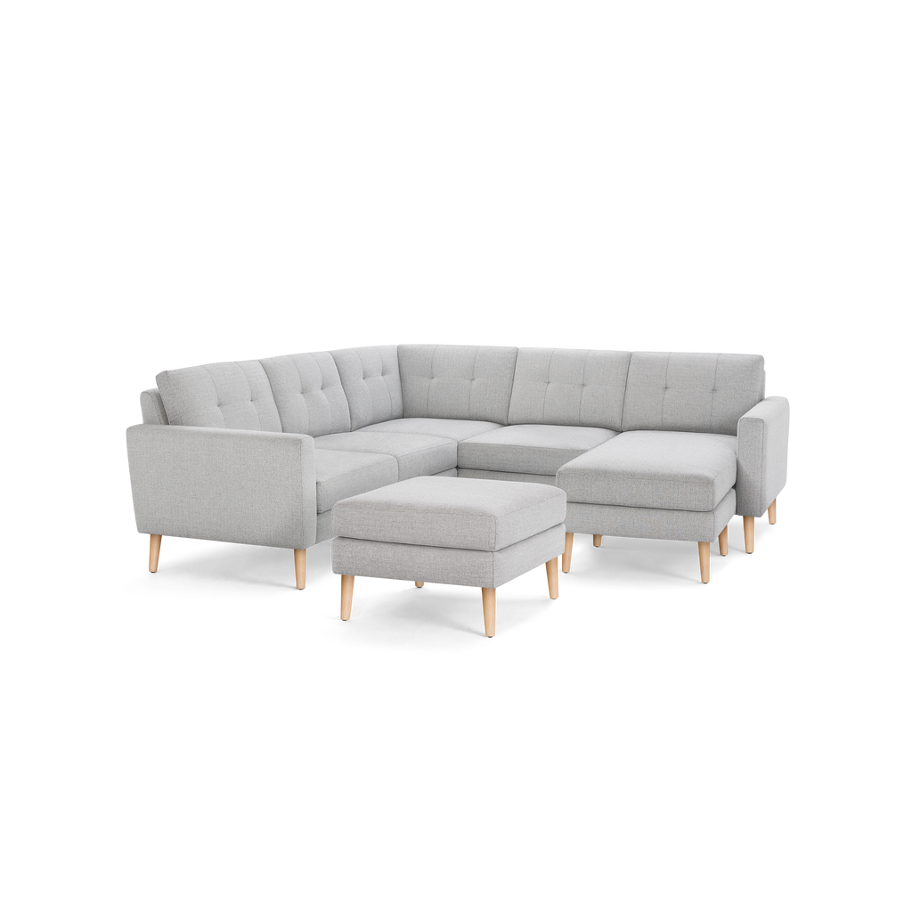 Light gray fabric sectional sofa with tufted back cushions, wooden legs, and matching ottoman, isolated on black background.