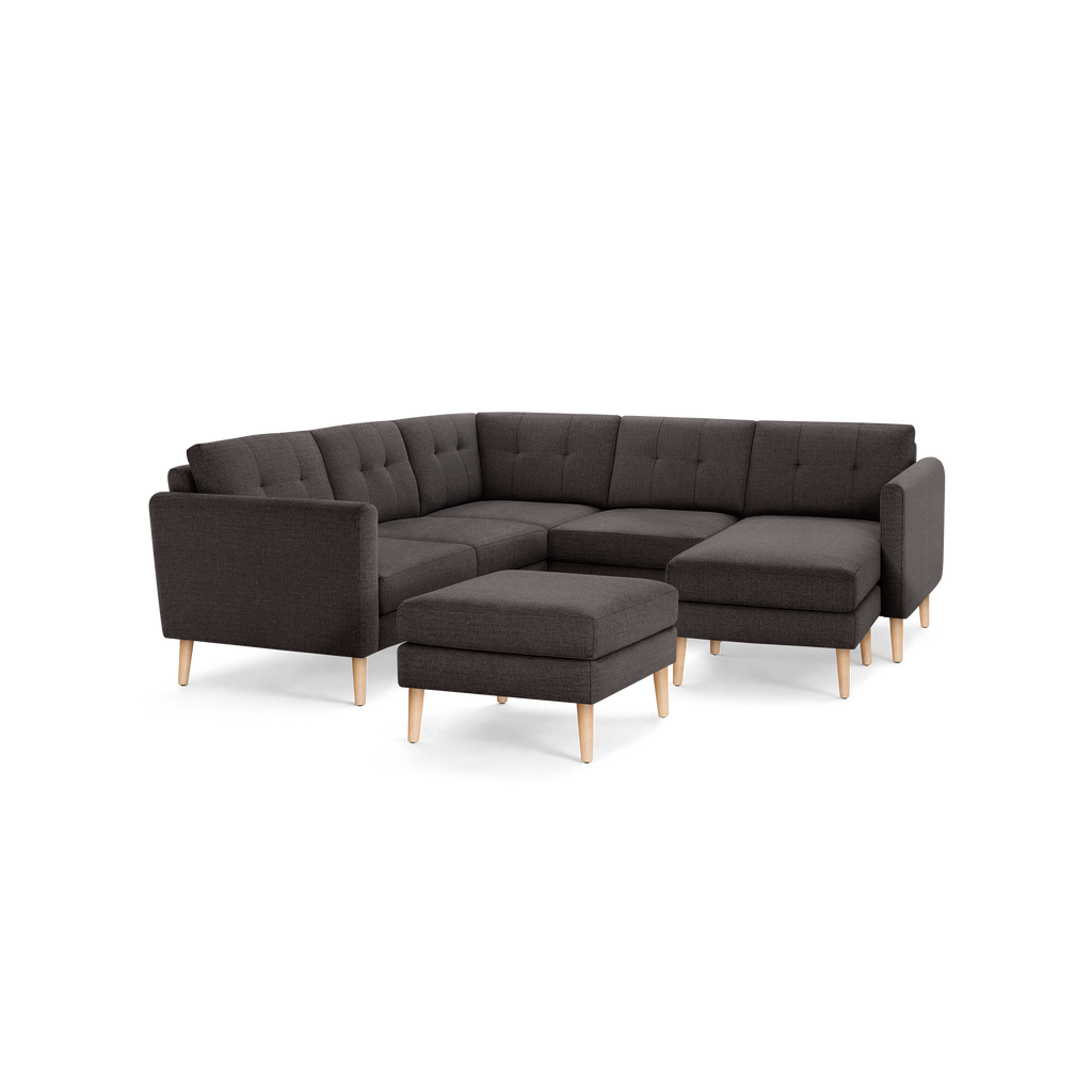 Dark gray fabric L-shaped sectional sofa with tufted back cushions, wooden legs, and matching ottoman isolated on black background.