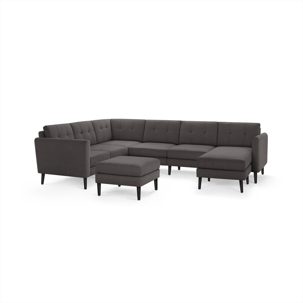 Dark gray modern sectional sofa with tufted cushions, black angled legs, and matching ottoman isolated on white background.