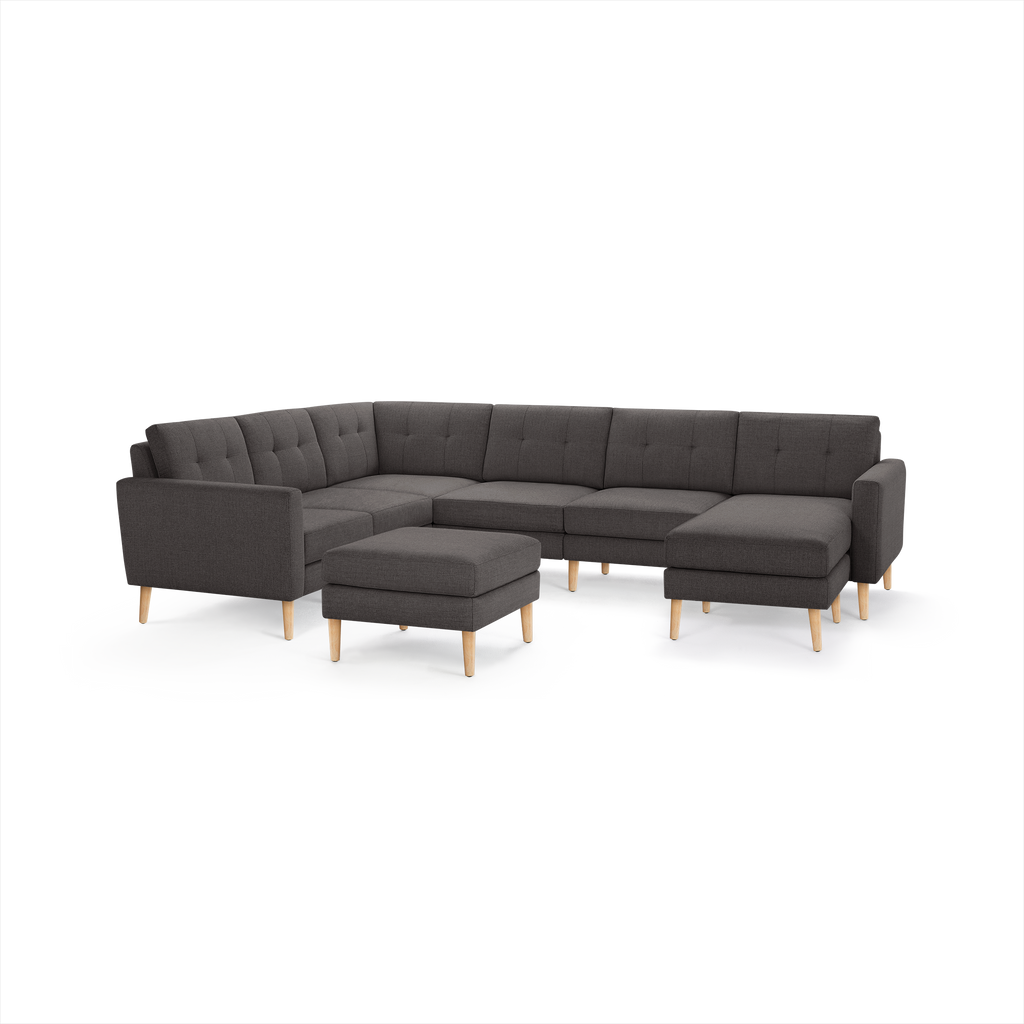 Modern dark gray fabric sectional sofa with tufted cushions, light wooden legs, and a matching ottoman on white background.