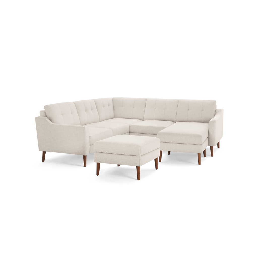 Light beige L-shaped sectional sofa with tufted back cushions, wooden legs, and matching ottoman on a black background.