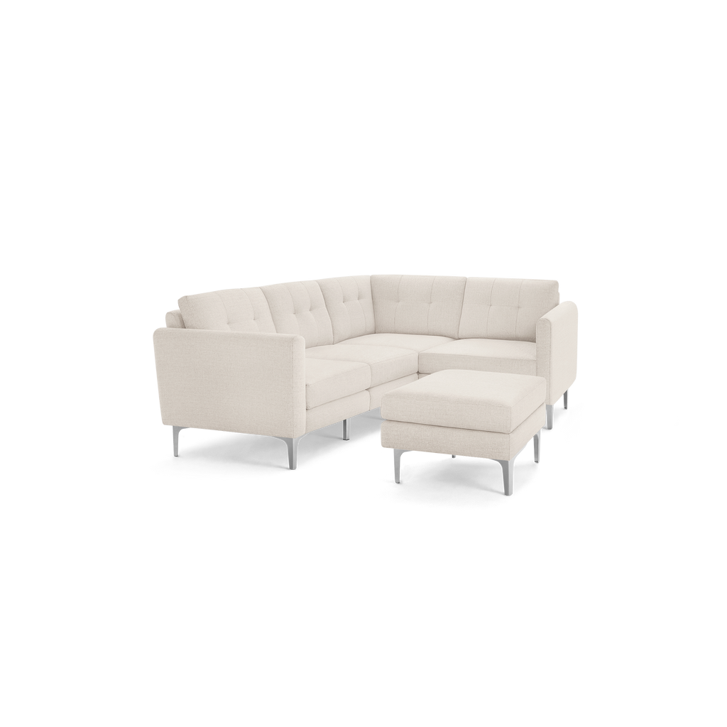 Beige fabric L-shaped sectional sofa with tufted back cushions and matching ottoman on metal legs, isolated on black background.