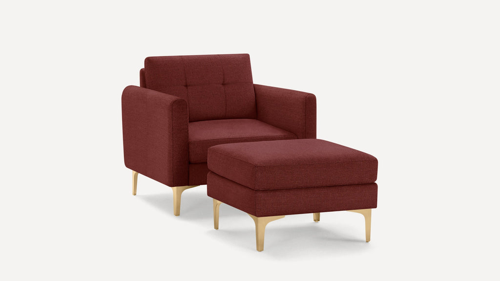 Modern burgundy fabric armchair with matching ottoman, featuring tufted backrest and gold-toned metal legs on white background.