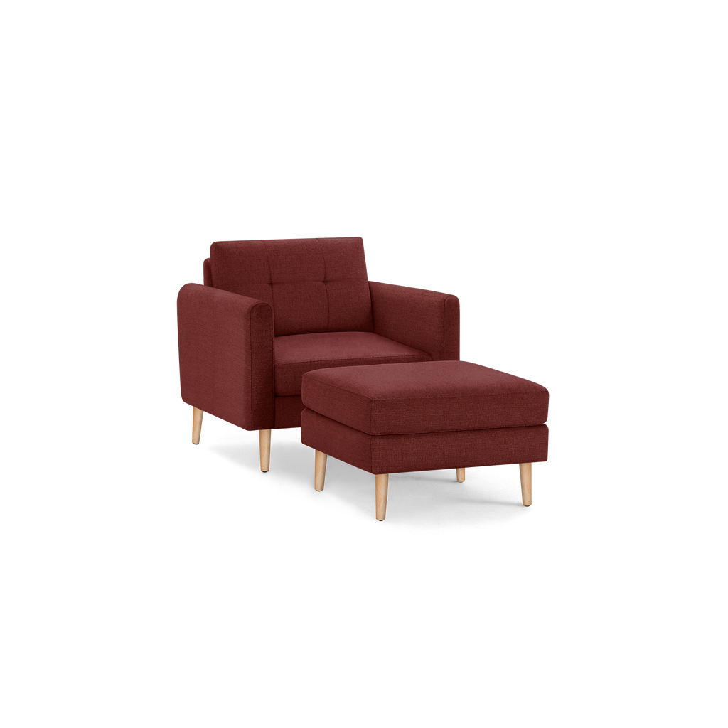 Modern red fabric armchair with matching ottoman, tufted backrest, and natural wooden legs, isolated on black background.