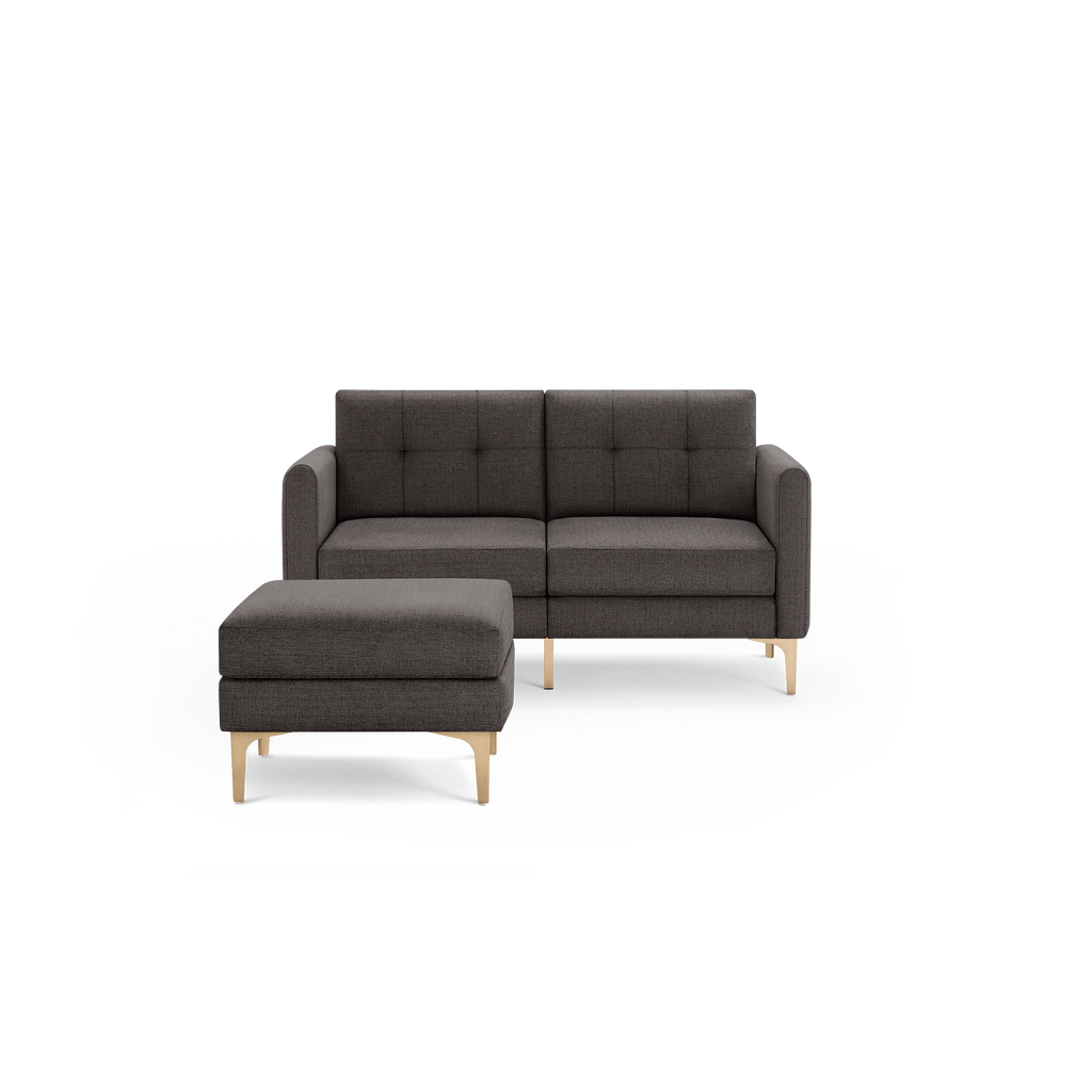 Modern dark gray two-seater sofa with matching ottoman, featuring tufted cushions and gold metal legs, isolated on black background.