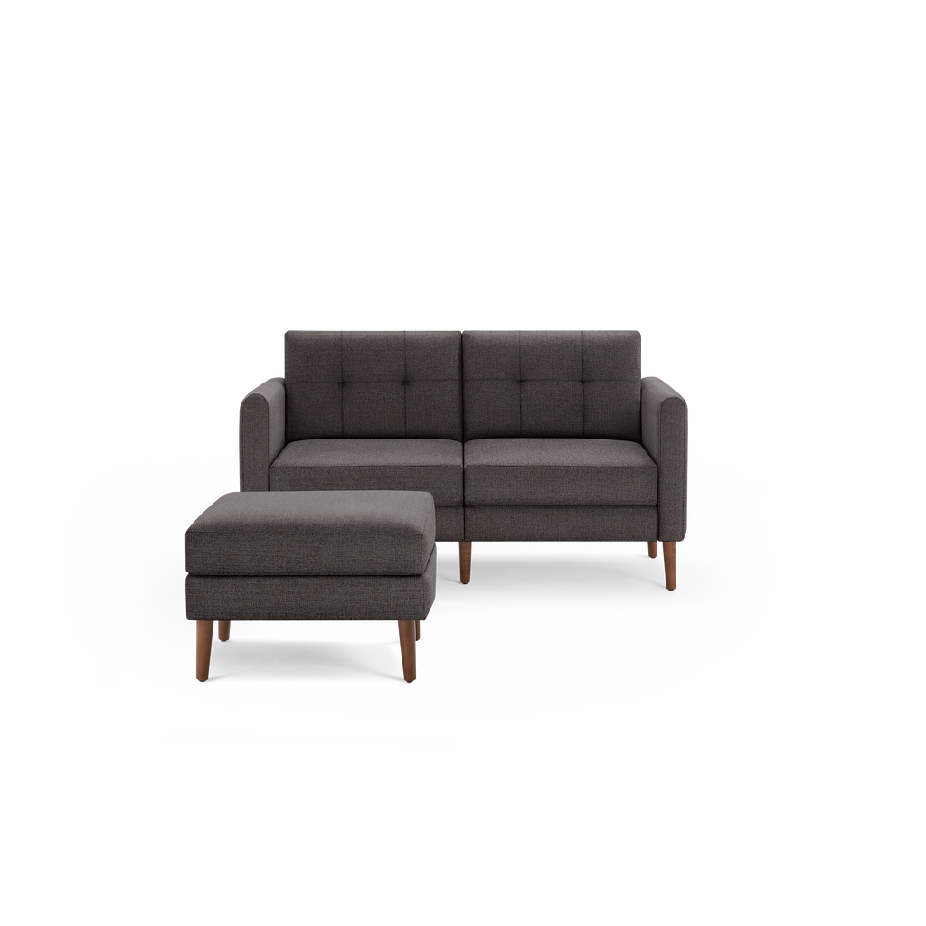 Dark gray upholstered modern sofa with matching ottoman, wooden legs, and tufted back cushions, isolated on white background.