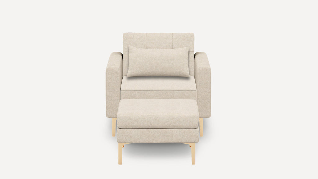 Beige upholstered armchair with matching rectangular lumbar pillow and ottoman, supported by light wooden legs, shown against a plain white background.