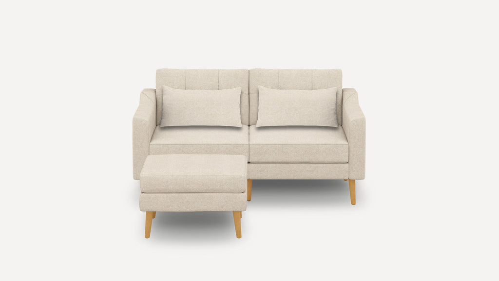 Beige fabric loveseat sofa with two matching pillows and a separate ottoman, featuring wooden legs and a modern design.