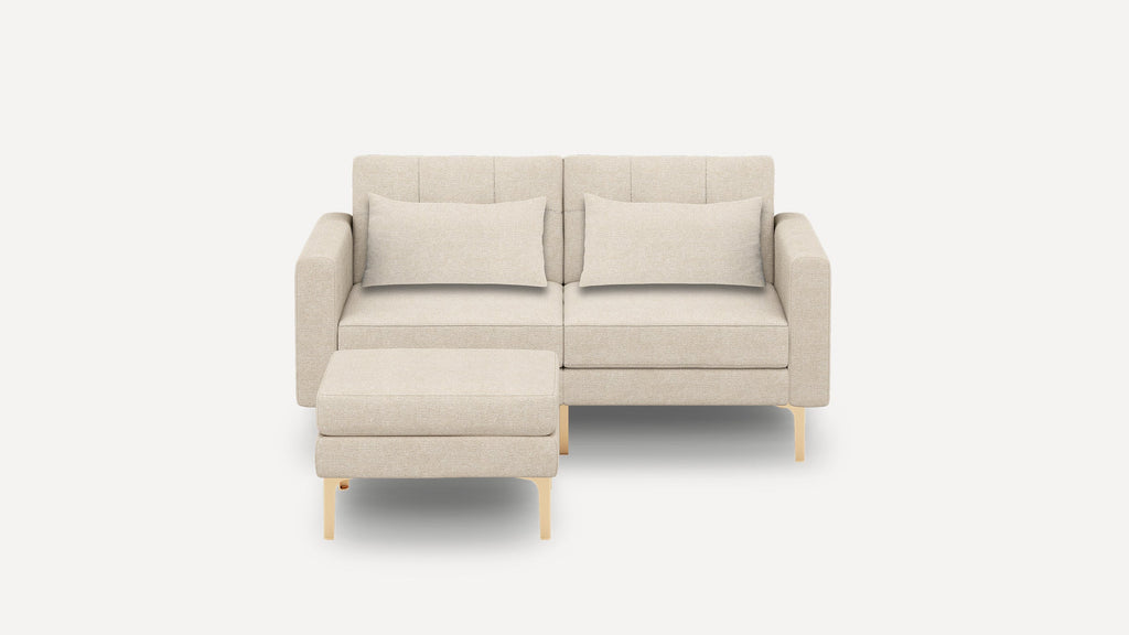 Beige modern two-seater sofa with matching ottoman, cushioned seats, back pillows, and light wooden legs on a white background.