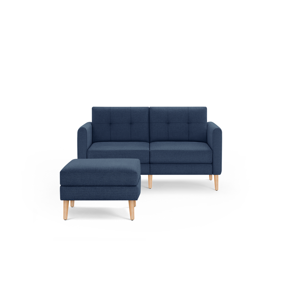 Navy blue modern two-seater sofa with matching ottoman, tufted back cushions, and light wooden legs on black background.