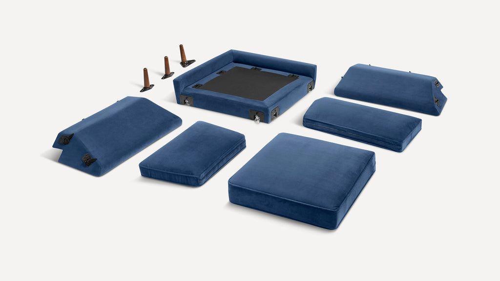 Disassembled blue velvet sofa cushions and frame pieces laid out with four brown wooden legs on a white background.