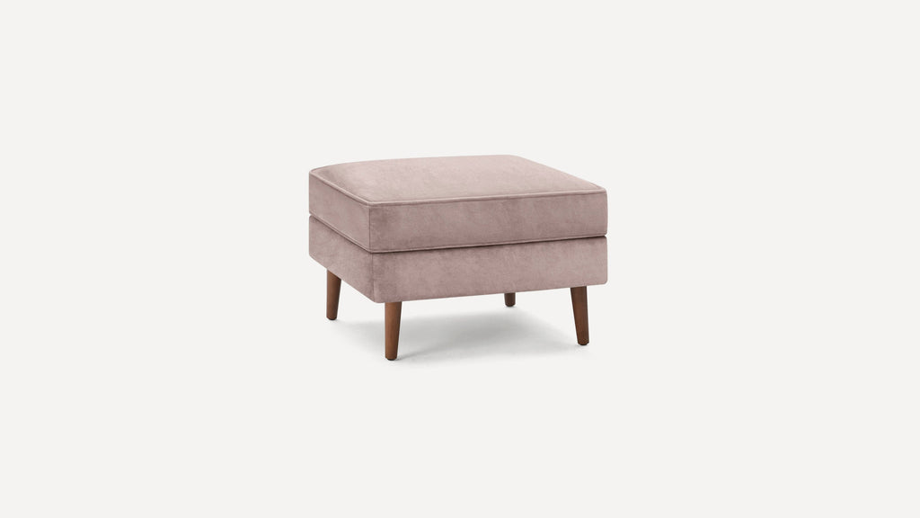 Square ottoman with plush pink velvet cushion and four wooden tapered legs on white background.
