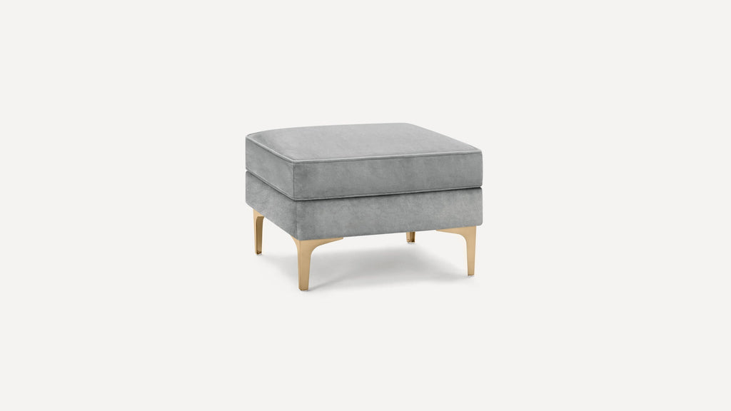 Square gray upholstered ottoman with gold metal legs on a white background, modern and minimalist design.