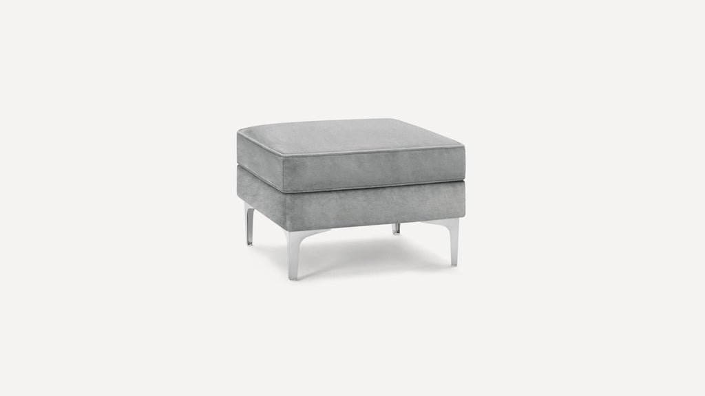 Modern square gray velvet ottoman with cushioned top and sleek silver metal legs, isolated on white background.