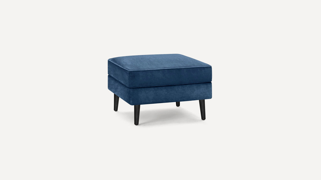 Square blue velvet ottoman with black tapered wooden legs, featuring a thick cushioned top for seating or resting feet.