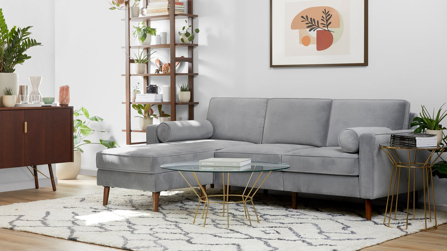 Modern living room with a light gray sectional sofa, round glass coffee table with gold base, wooden shelf, and mid-century cabinet.