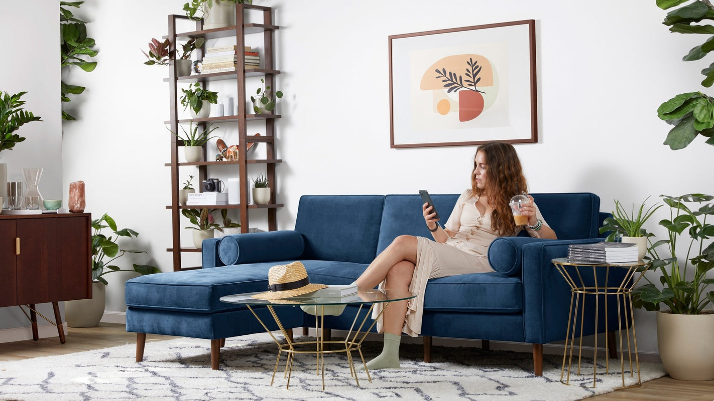 Woman sitting on navy blue velvet sectional sofa with chaise, in modern living room with plants and gold-accented tables.