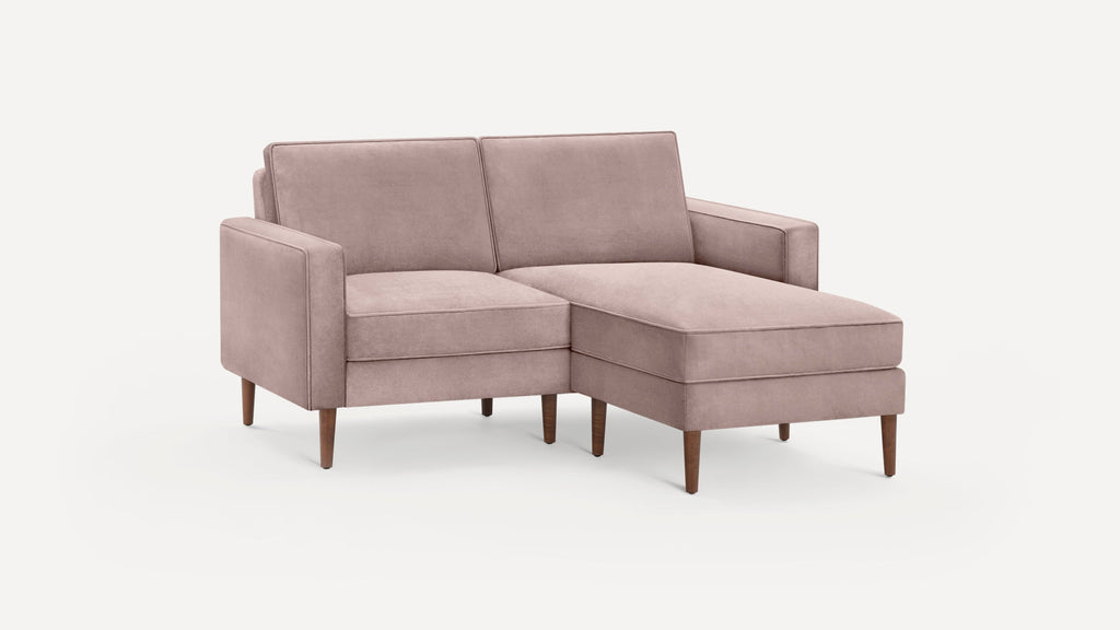 Modern blush pink velvet sectional sofa with wooden legs, featuring a chaise lounge on the right side, set against a plain white background.
