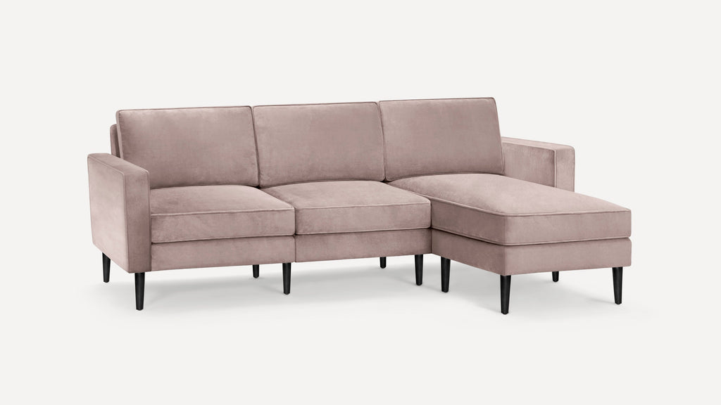 Blush pink velvet sectional sofa with three seat cushions, three back cushions, slim black legs, and a right chaise lounge.