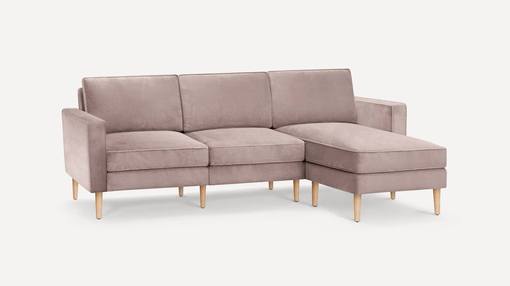 Modern L-shaped sofa in soft blush pink velvet with wooden legs, featuring a chaise lounge on the right side.
