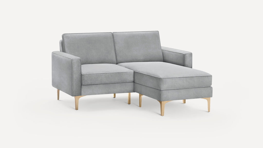Light gray velvet sectional sofa with chaise lounge and gold metal legs on a plain white background.