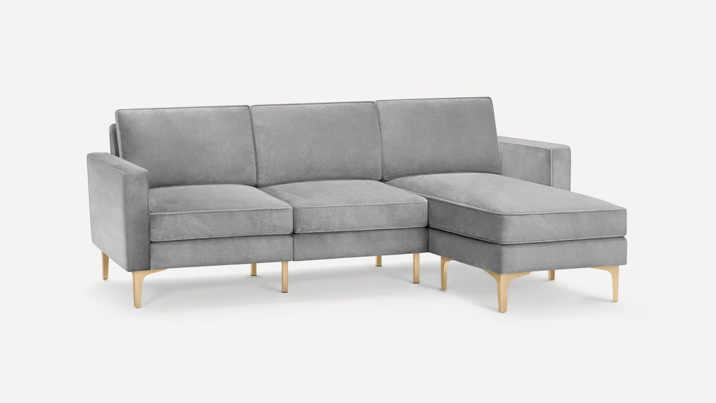 Light gray velvet L-shaped sectional sofa with gold metal legs and three back cushions on a white background.