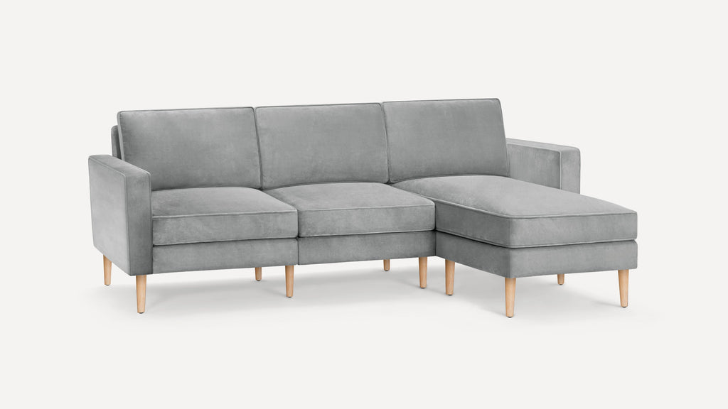 Modern light gray sectional sofa with wooden legs, featuring three seat cushions and a chaise lounge on the right side.