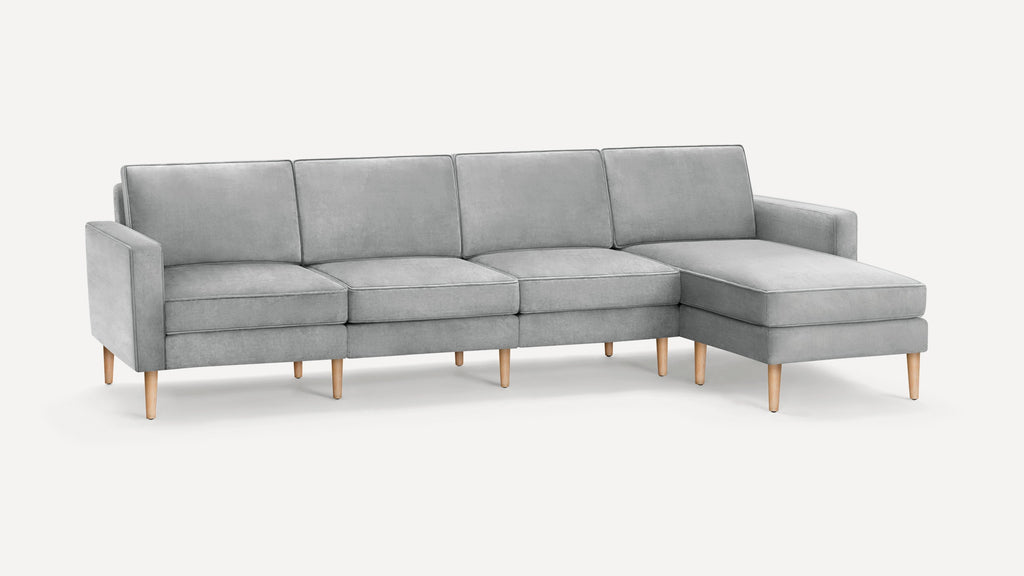 Light gray sectional sofa with four seat cushions, wooden legs, and a chaise lounge on the right side, set against a plain background.