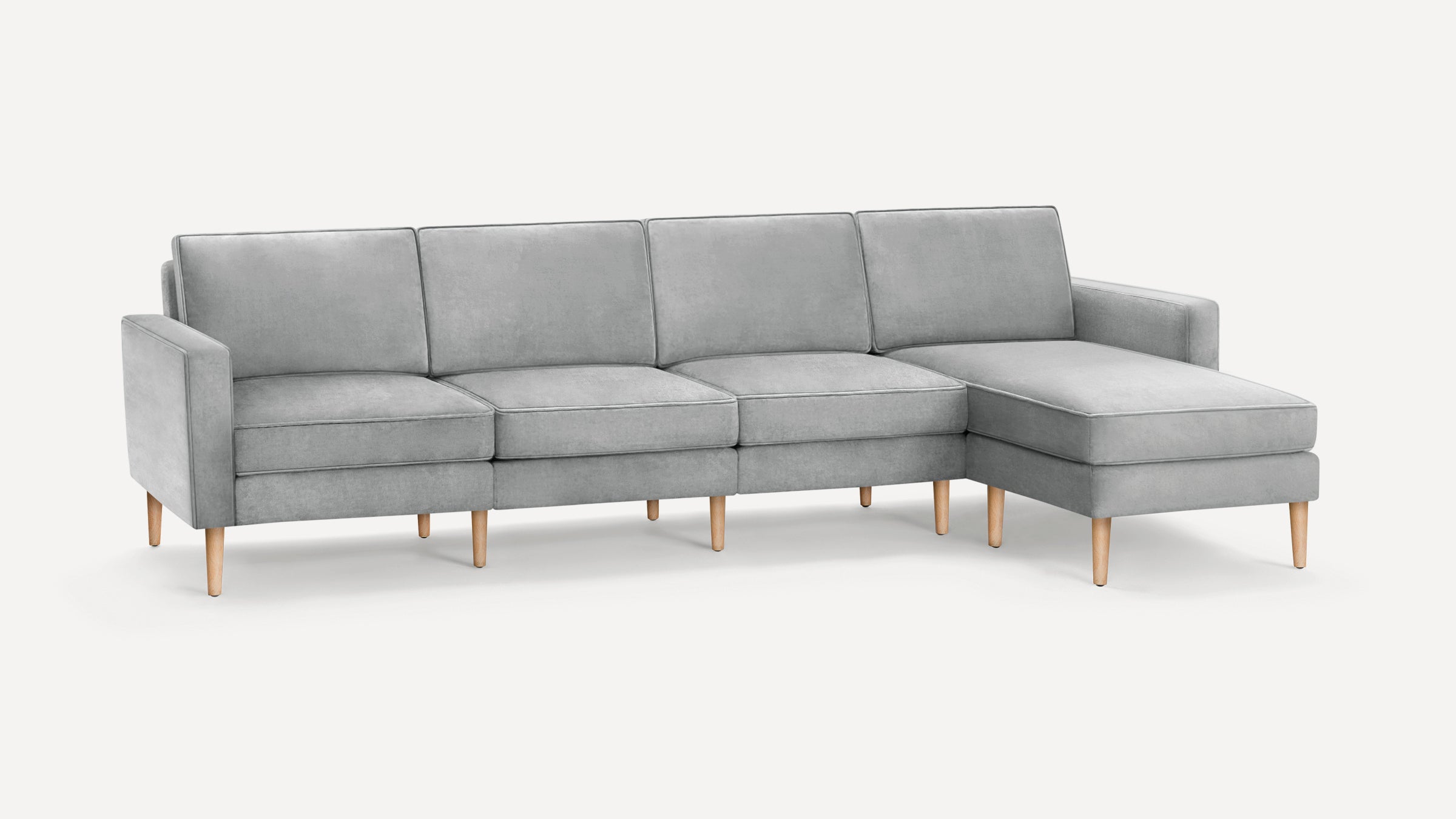 Light gray sectional sofa with four seat cushions, wooden legs, and a chaise lounge on the right side, set against a plain background.