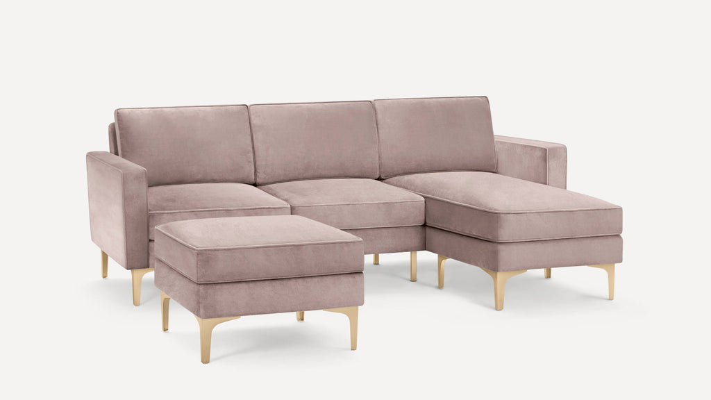 Modern dusty pink velvet sectional sofa with three seat cushions, matching ottoman, and gold metal legs on white background.