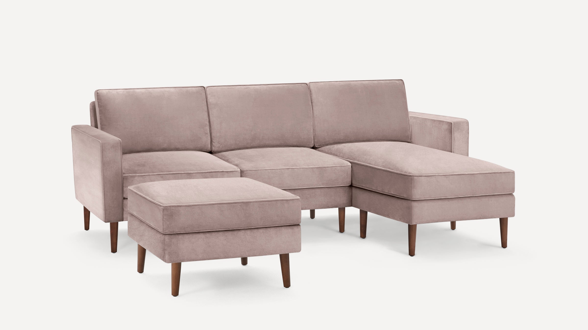 Modern blush pink sectional sofa with chaise lounge and matching ottoman, featuring wooden tapered legs and plush cushions.