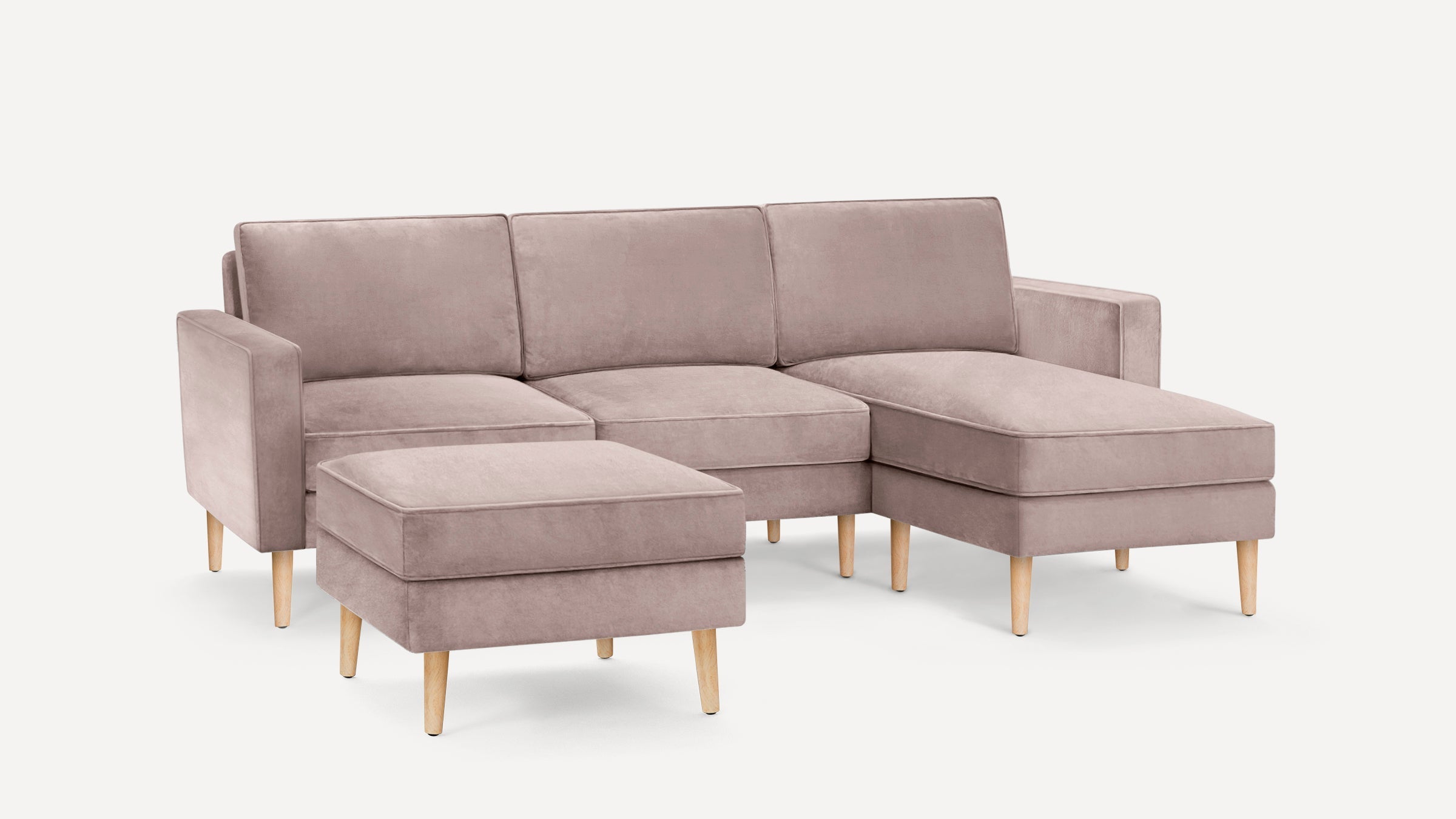 Modern L-shaped sectional sofa with matching ottoman, upholstered in taupe velvet fabric, supported by light wooden legs.