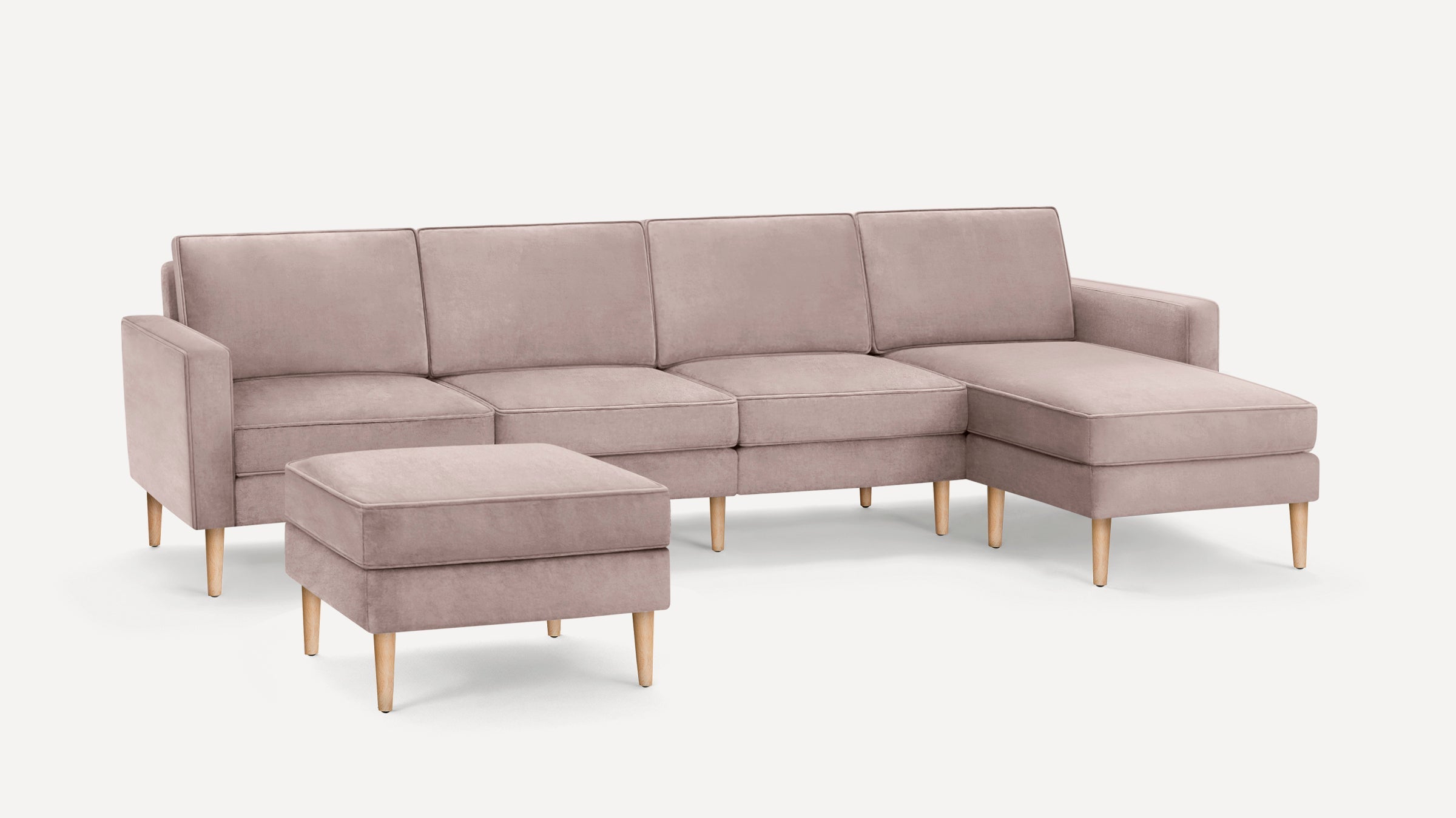 Light pink velvet sectional sofa with wooden legs and matching ottoman on white background.