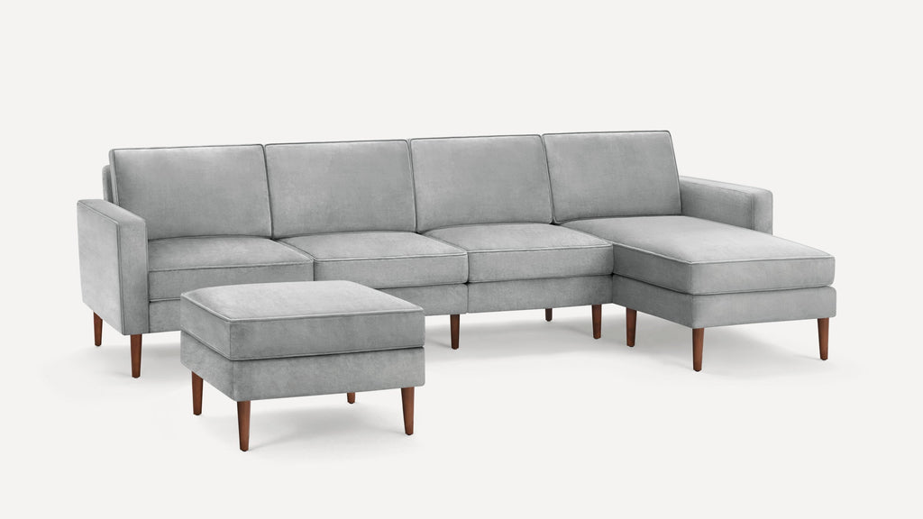 Light gray sectional sofa with four seat cushions, a matching ottoman, and tapered wooden legs on a white background.