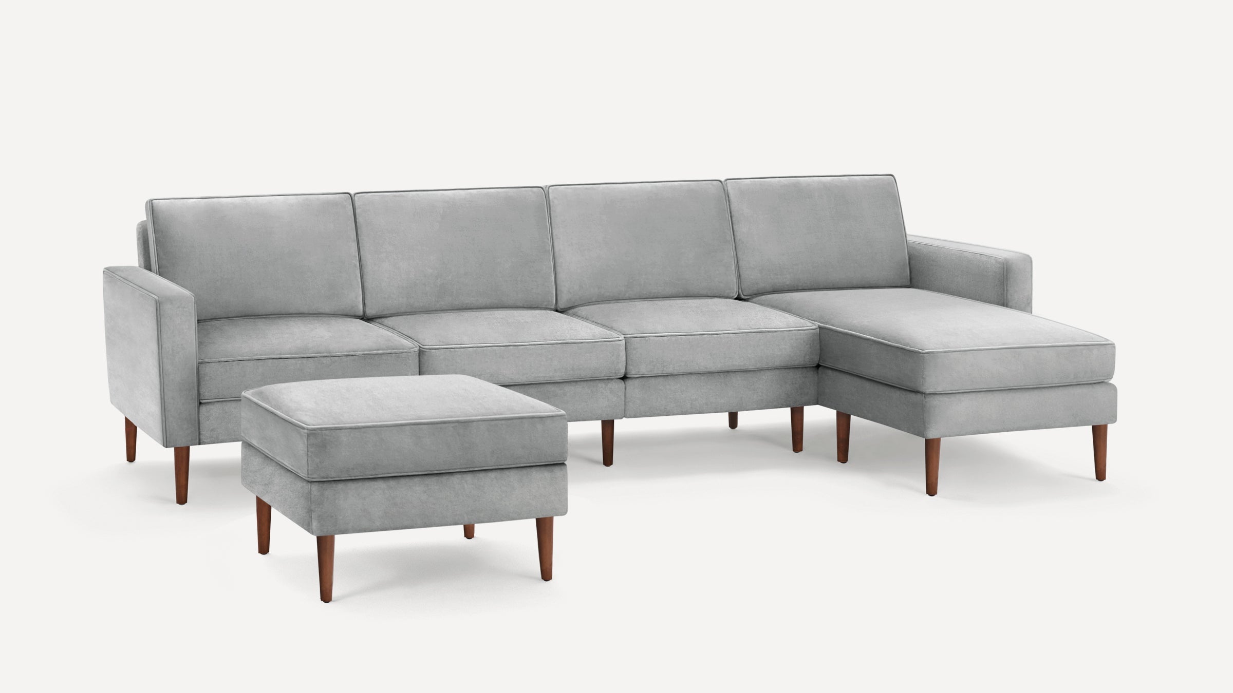 Light gray sectional sofa with four seat cushions, a matching ottoman, and tapered wooden legs on a white background.