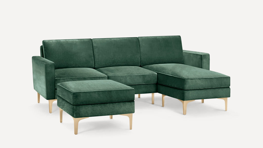 Modern green velvet sectional sofa with chaise lounge and matching ottoman on light wooden legs, set against plain white background.