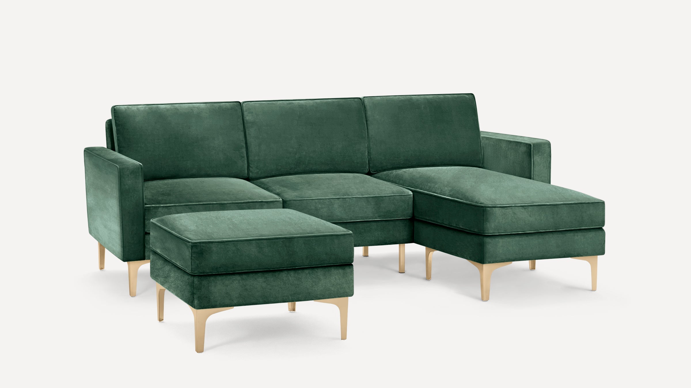 Modern green velvet sectional sofa with chaise lounge and matching ottoman on light wooden legs, set against plain white background.