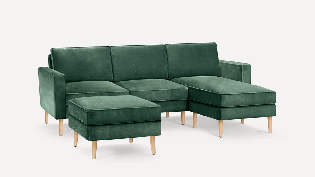 Dark green velvet sectional sofa with wooden legs, including a matching ottoman, set against a white background.