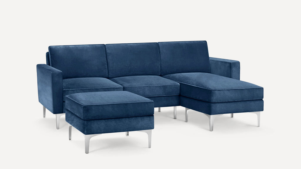 Modern navy blue velvet sectional sofa with three seat cushions, chaise lounge, and matching ottoman, supported by sleek silver metal legs.