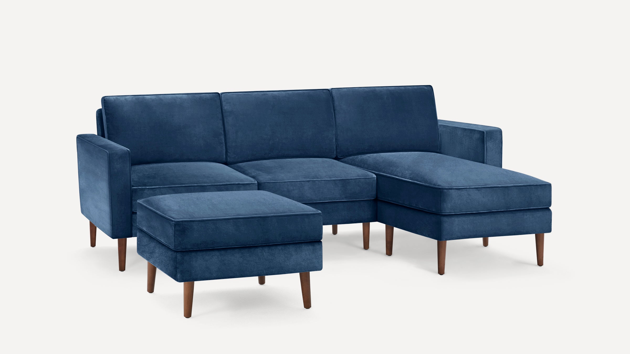 Modern navy blue velvet sectional sofa with wooden legs and matching ottoman on white background.