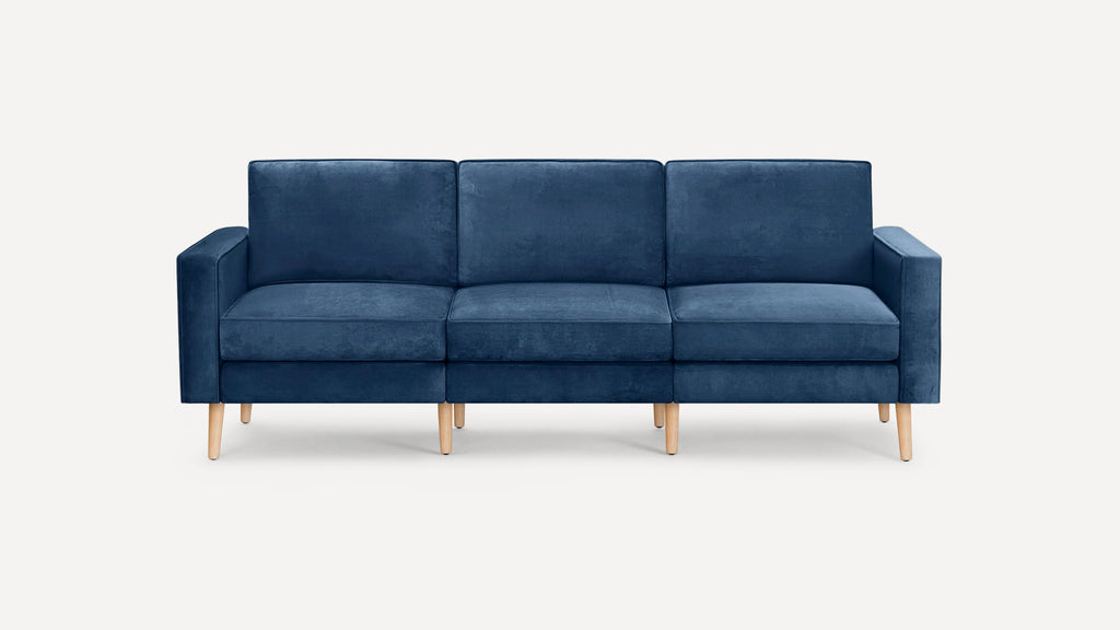 Modern three-seater sofa upholstered in deep blue velvet fabric with light wooden tapered legs and square armrests, set against a plain white background.