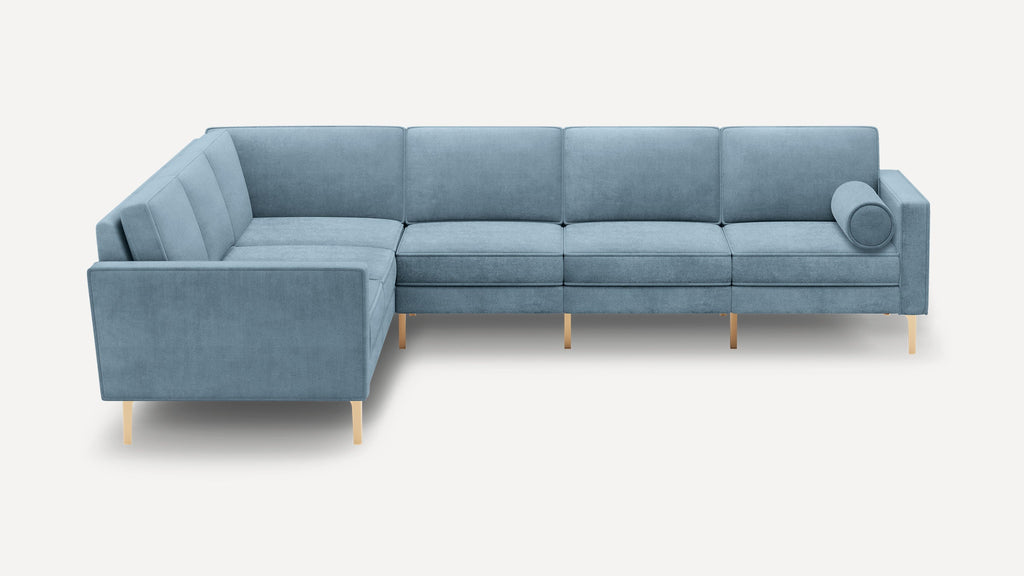 Modern L-shaped sectional sofa upholstered in light blue fabric with straight arms, tufted cushions, brass legs, and a round bolster pillow.